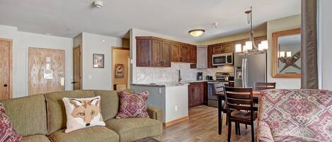 A spacious living room with a sofa and coffee table, a dining area with chairs, and a kitchen with wooden cabinets and appliances.