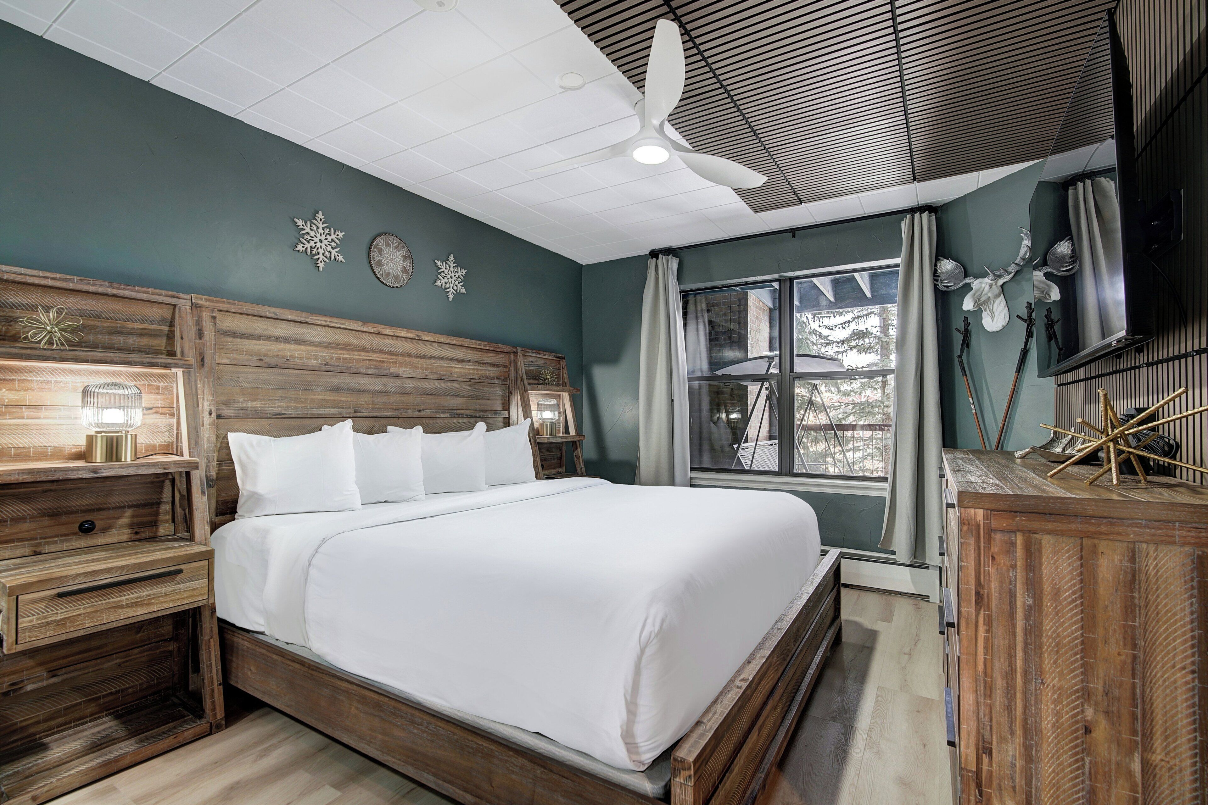 Une chambre à coucher avec un lit en bois, des tables de chevet, un ventilateur de plafond et une grande fenêtre avec des rideaux.