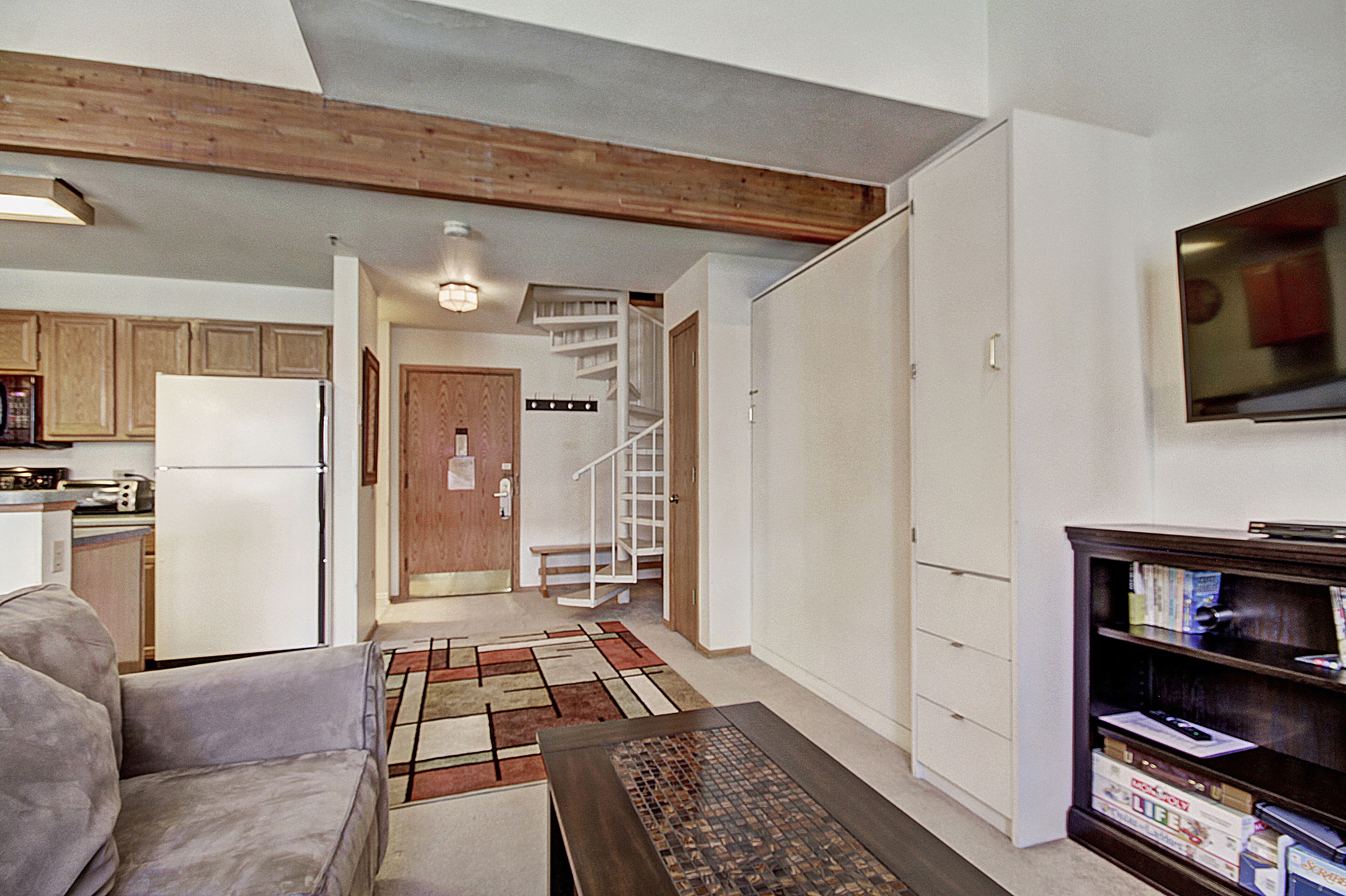 A compact living space with a kitchenette, a sofa, a coffee table, a bookshelf, and a staircase leading to an upper level.