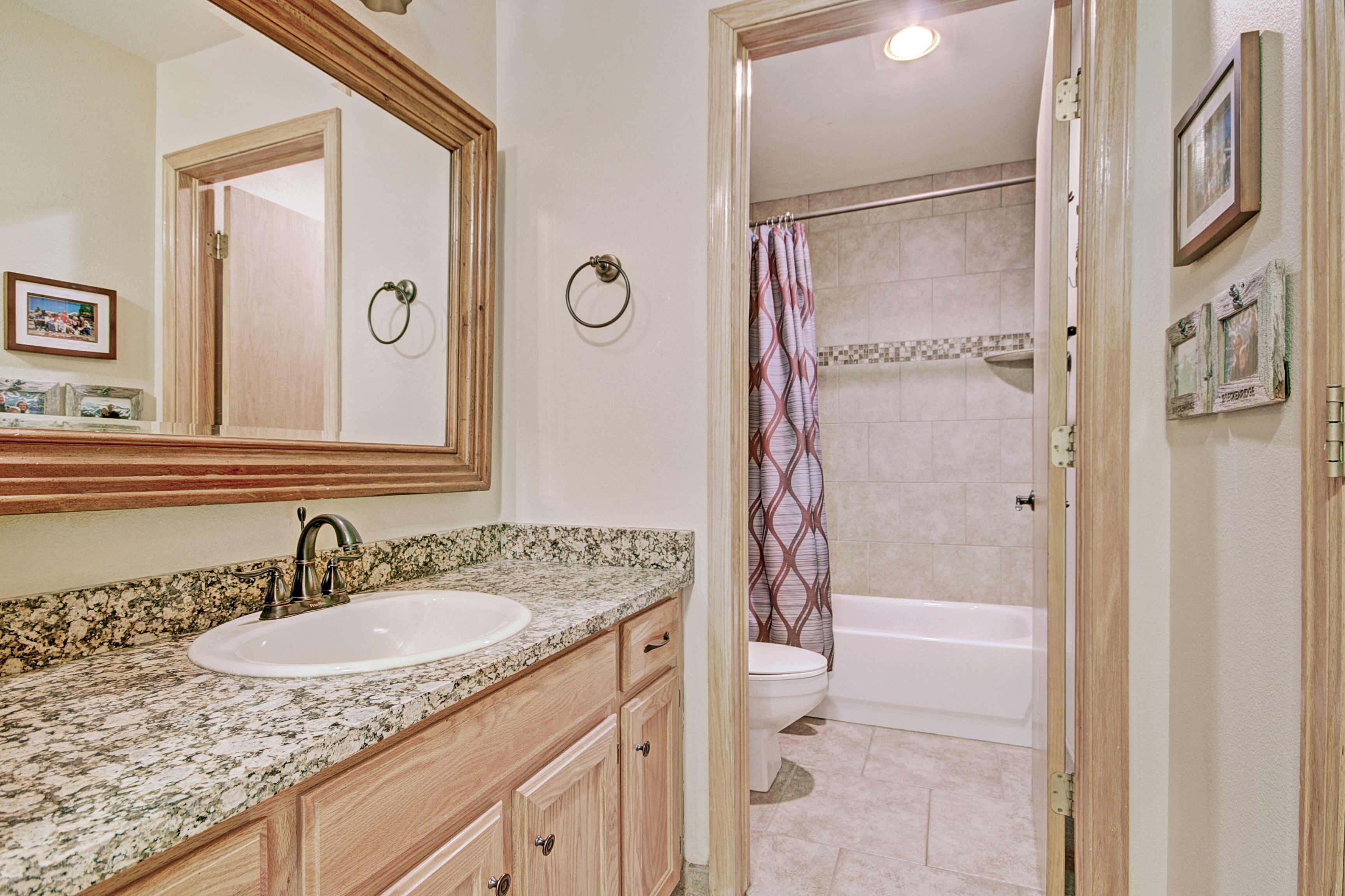 Baño con lavamanos de encimera de granito, armarios de madera, un espejo grande, inodoro y ducha con cortina estampada.