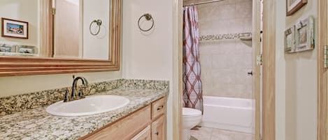 A bathroom with a granite countertop sink, wooden cabinets, a large mirror, a toilet, and a shower with a patterned curtain.
