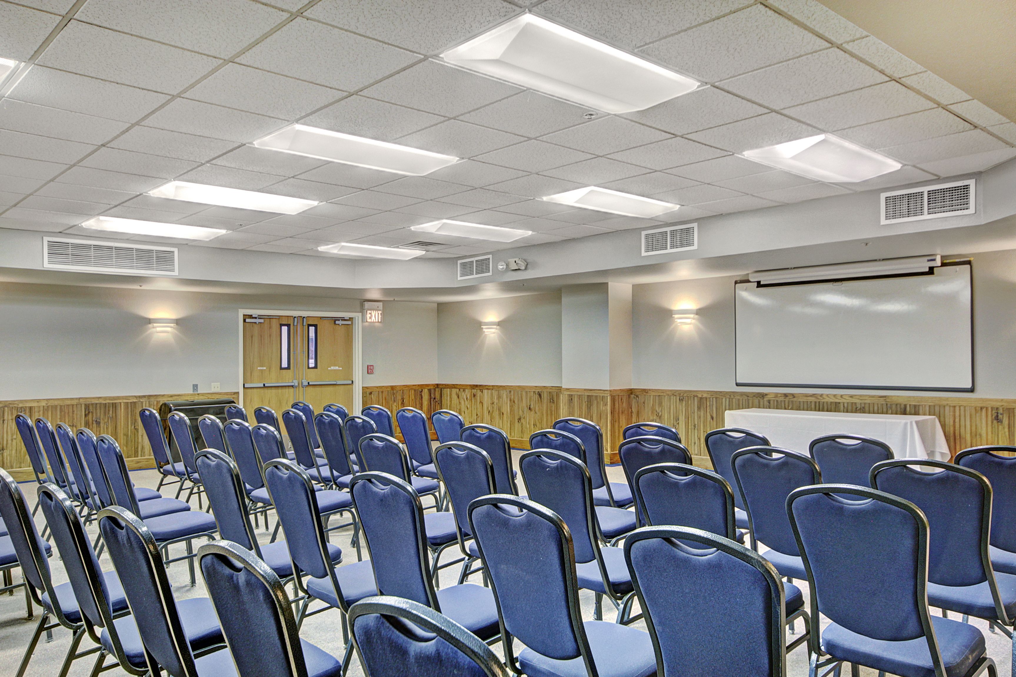A conference room with rows of blue chairs, a white projection screen, and a wooden panel wall.