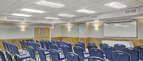 A conference room with rows of blue chairs, a white projection screen, and a wooden panel wall.