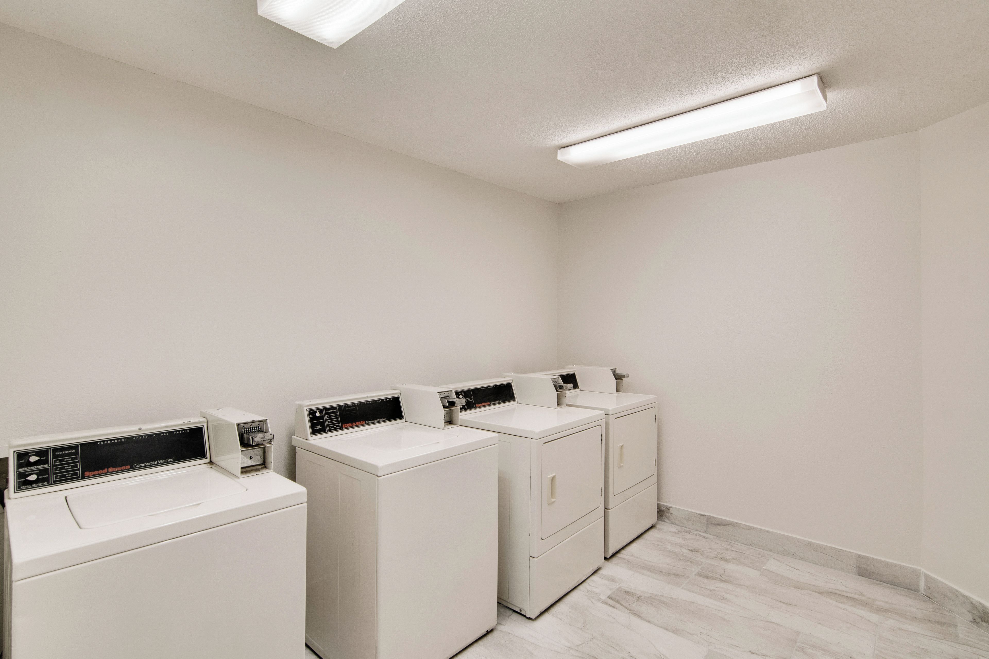 A laundry facility with four white washing machines and dryers, a tiled floor, and a plain white wall.