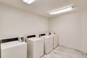Une buanderie équipée de quatre machines à laver et de quatre sèche-linge blancs, d’un sol carrelé et d’un mur blanc uni.