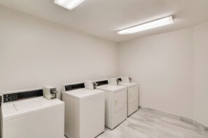 Une buanderie équipée de quatre machines à laver et de quatre sèche-linge blancs, d’un sol carrelé et d’un mur blanc uni.