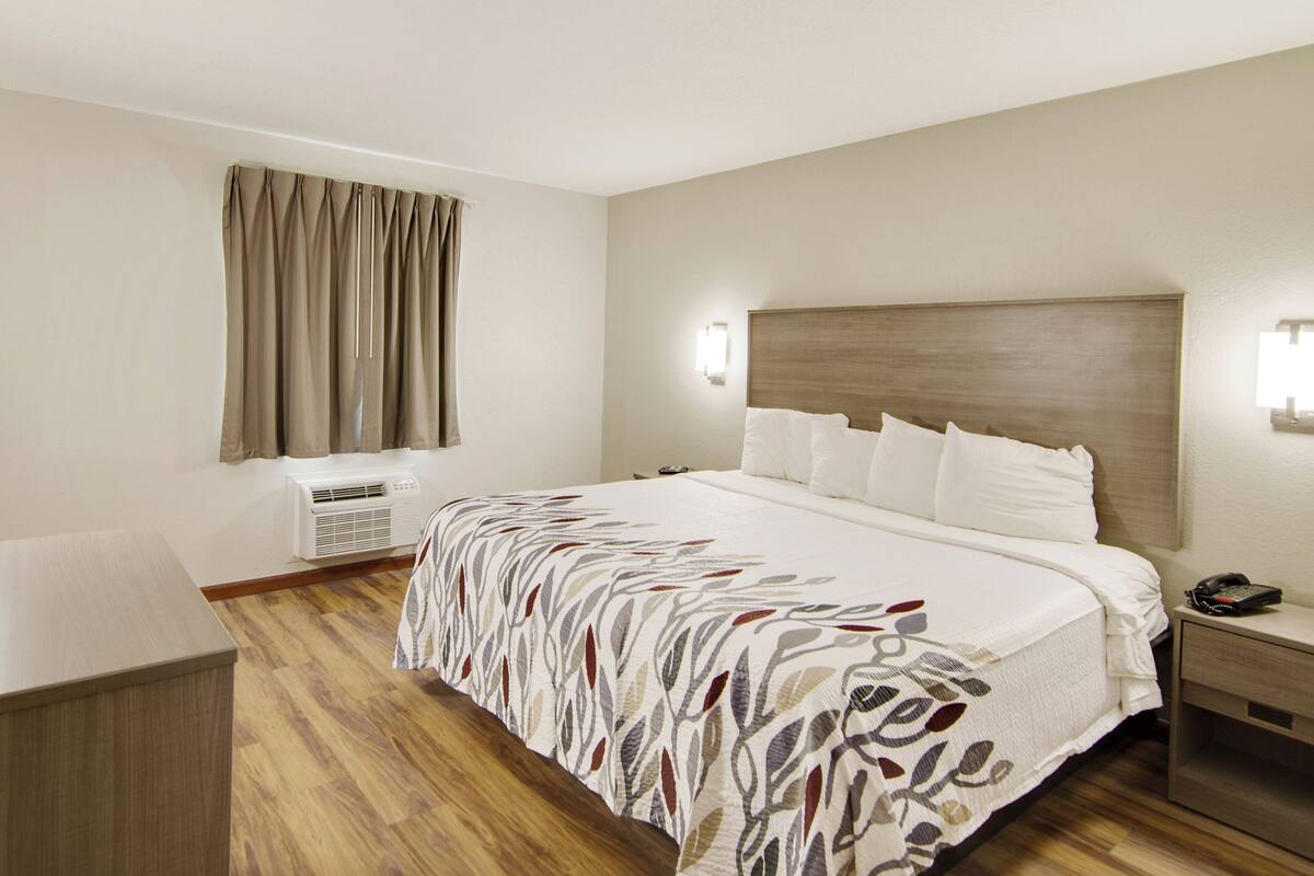Een hotelkamer met een groot bed, een nachtkastje met een lamp, een wandairconditioner en een raam met gordijnen.