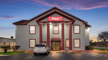 A hotel with a red and white facade, a central entrance with columns, and a parking lot in front.