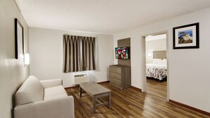 A hotel room with a wooden floor, a sofa, a small table, a TV, a bed, and a picture on the wall. - Red Roof Inn & Suites Pensacola - NAS Corry (Pensacola)