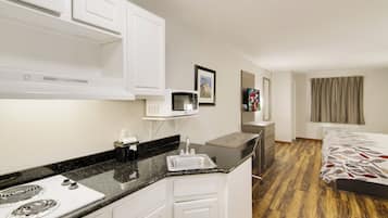 Habitación de hotel compacta con kitchenette, una cama con ropa de cama estampada, un televisor de pantalla plana y un ventanal con cortinas.