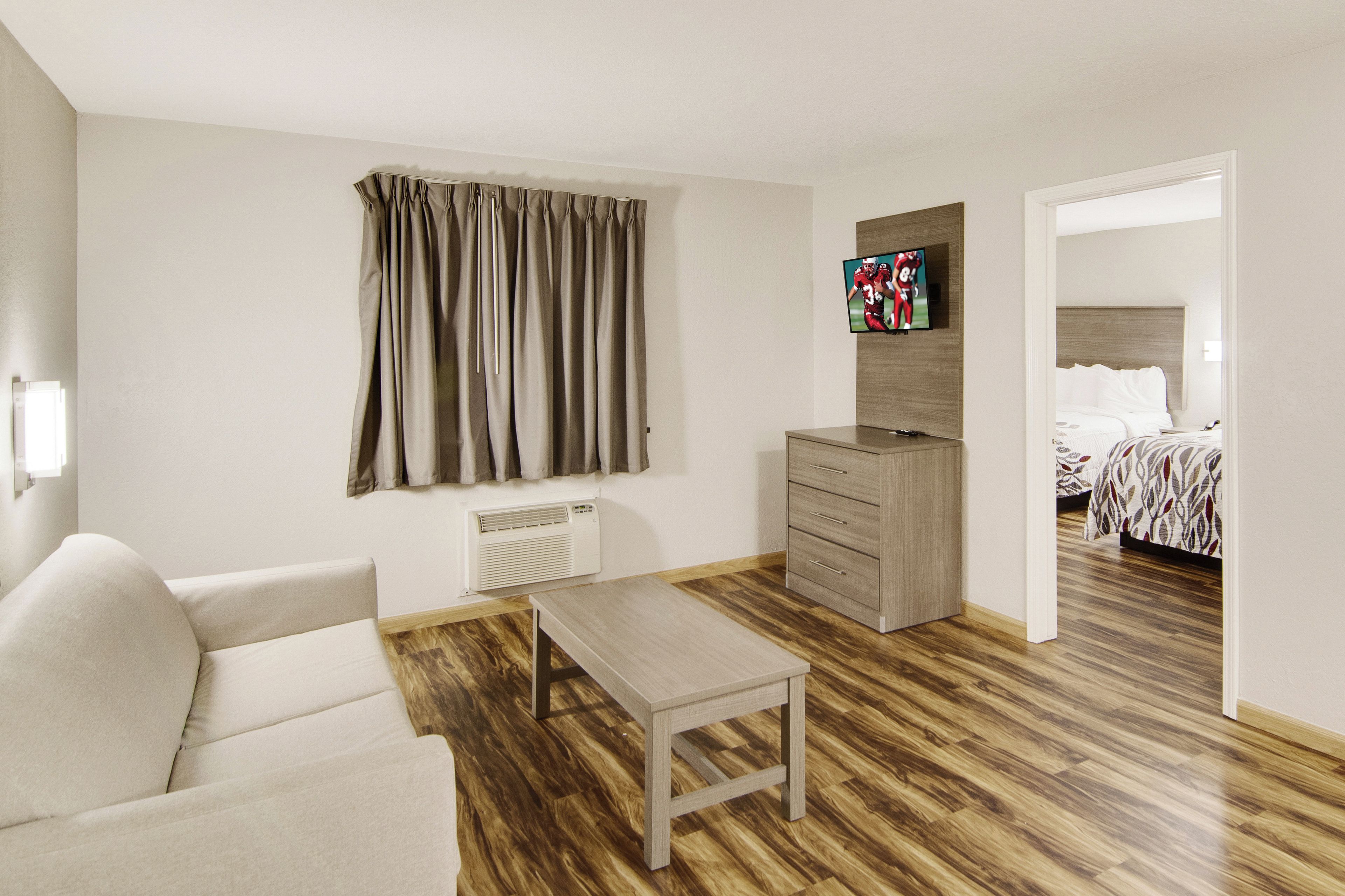 A hotel room with a wooden floor, a sofa, a small table, a dresser, a television mounted on the wall, and a bed with a patterned bedspread.