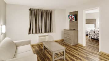 A hotel room with a wooden floor, a sofa, a small table, a dresser, a television mounted on the wall, and a bed with a patterned bedspread.