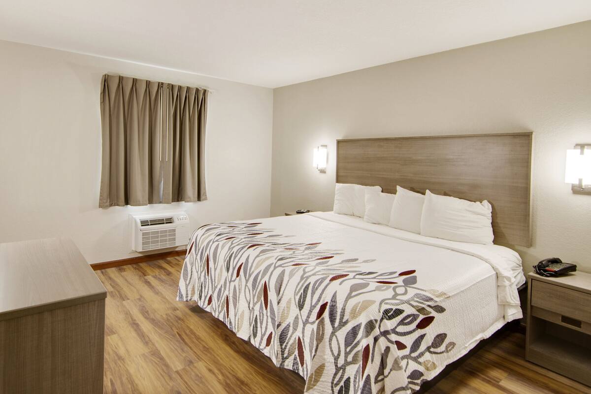 Habitación de hotel con una cama grande, una mesita de noche con lámpara, aire acondicionado en la pared y una ventana con cortinas.