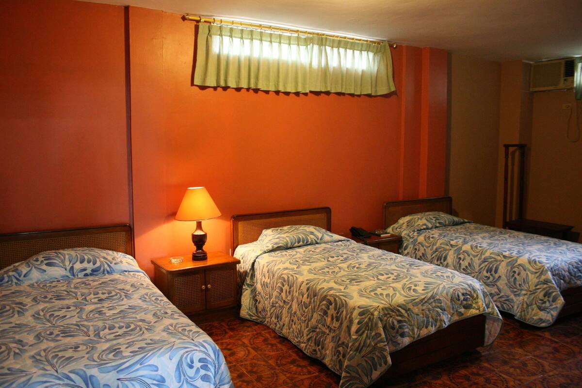 A hotel room with two single beds, a nightstand with a lamp, a window with a curtain, and an air conditioning unit.