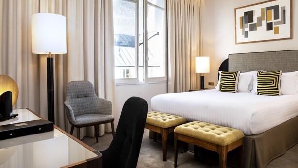 Una habitación de hotel con una cama, dos mesitas de noche, un escritorio, una silla y una ventana con cortinas.