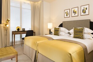 A hotel room with two beds, a desk, a chair, and a lamp.