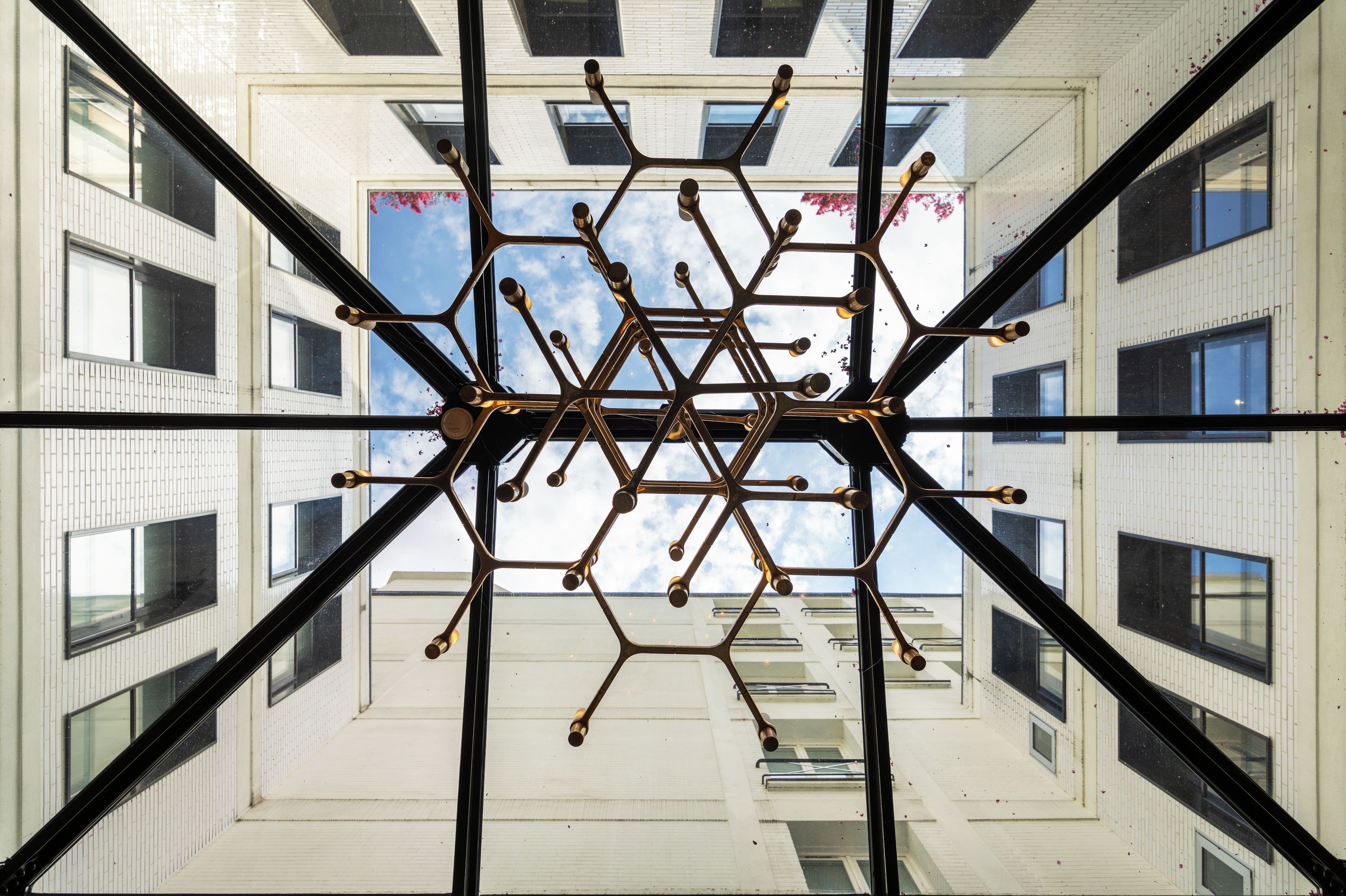 Una estructura geomètrica simètrica amb diversos braços i elements esfèrics, vista a través d'una superfície espeillada que reflecteix un edifici amb finestres i un cel blau.