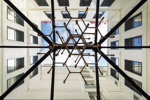 Een symmetrische, geometrische constructie met meerdere armen en bolvormige elementen, gezien door een spiegelend oppervlak dat een gebouw met ramen en een heldere lucht weerspiegelt.
