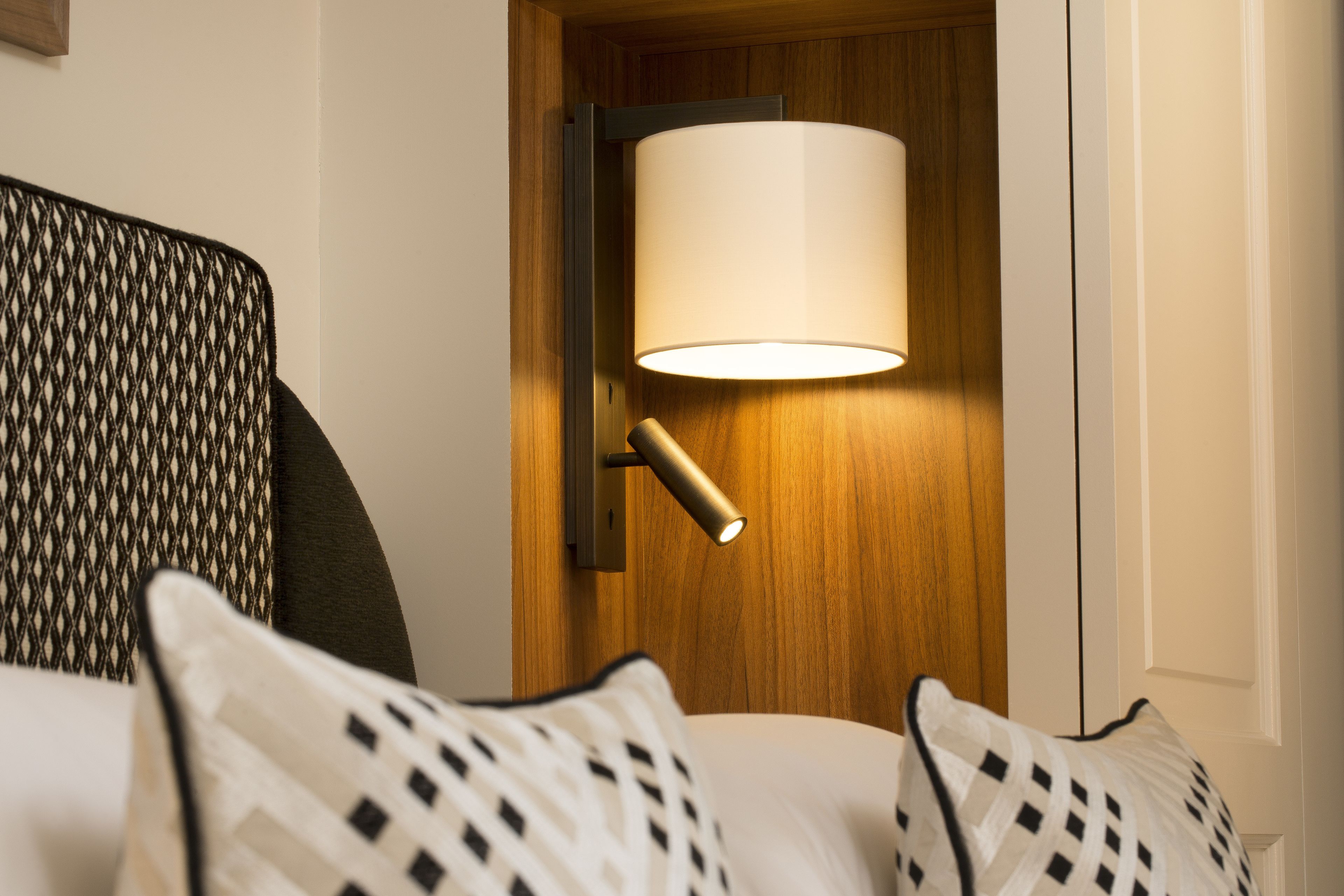 A hotel room with a wooden door, a wall-mounted lamp, and patterned pillows.