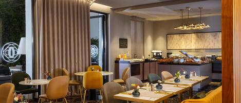 A modern hotel dining area with round tables, comfortable seating, a kitchen in the background, and a large window offering a view.