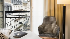 Habitación de hotel con una cama, un sillón estampado, una lámpara de pie y vistas a edificios a través de una ventana con barandilla de metal.