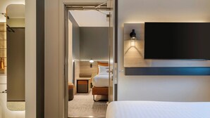 Une chambre d’hôtel avec un lit, un téléviseur fixé au mur, une porte donnant sur une salle de bain et une petite table avec une lampe.