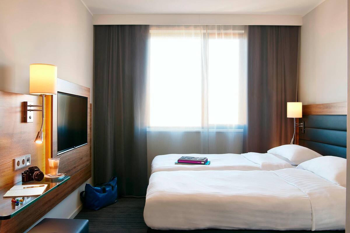 A hotel room with a bed, a desk with a lamp, a TV, and a window with curtains.