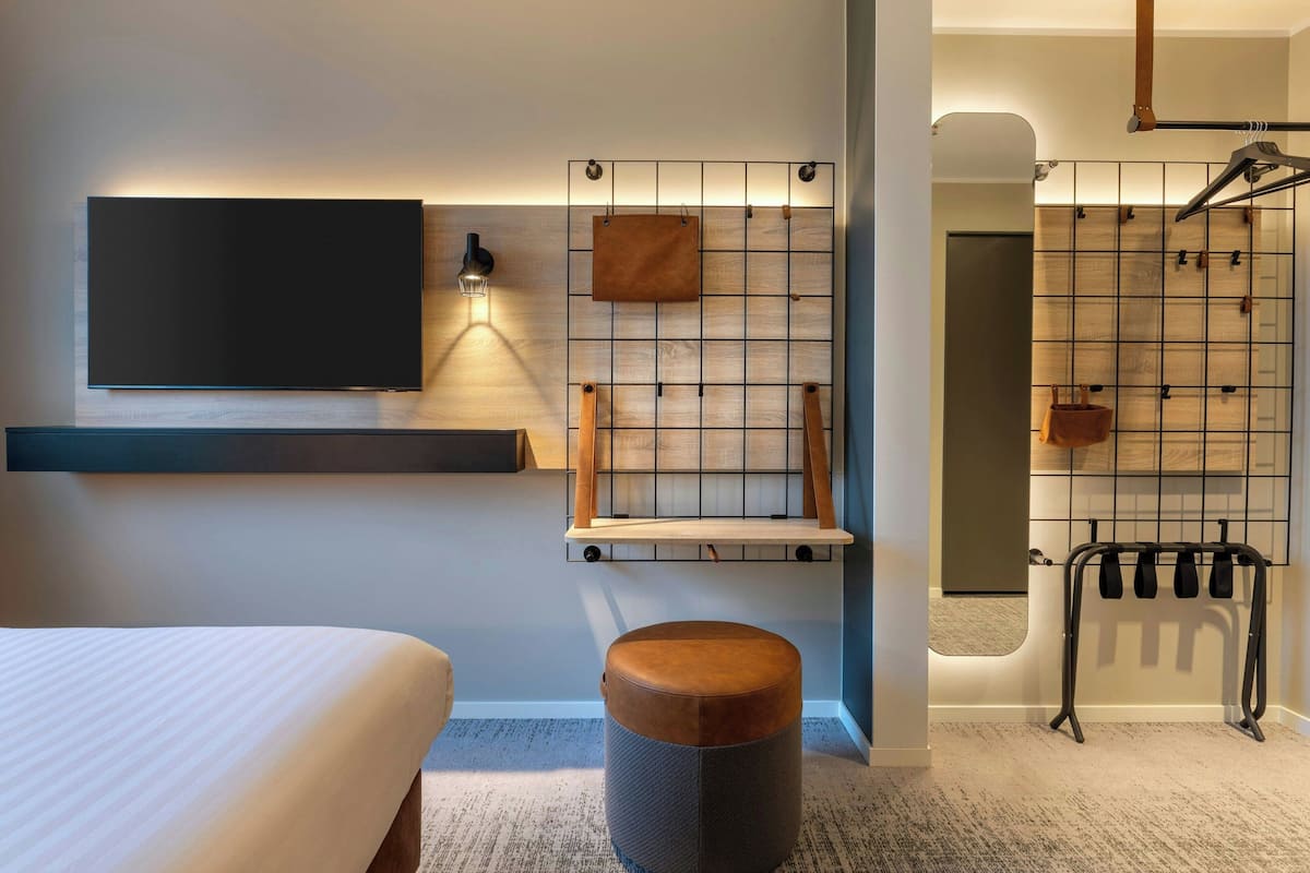 A modern hotel room with a large flat-screen TV, a wooden shelf, a hanging light, a grid-patterned wall, a towel rack, and a small round ottoman.