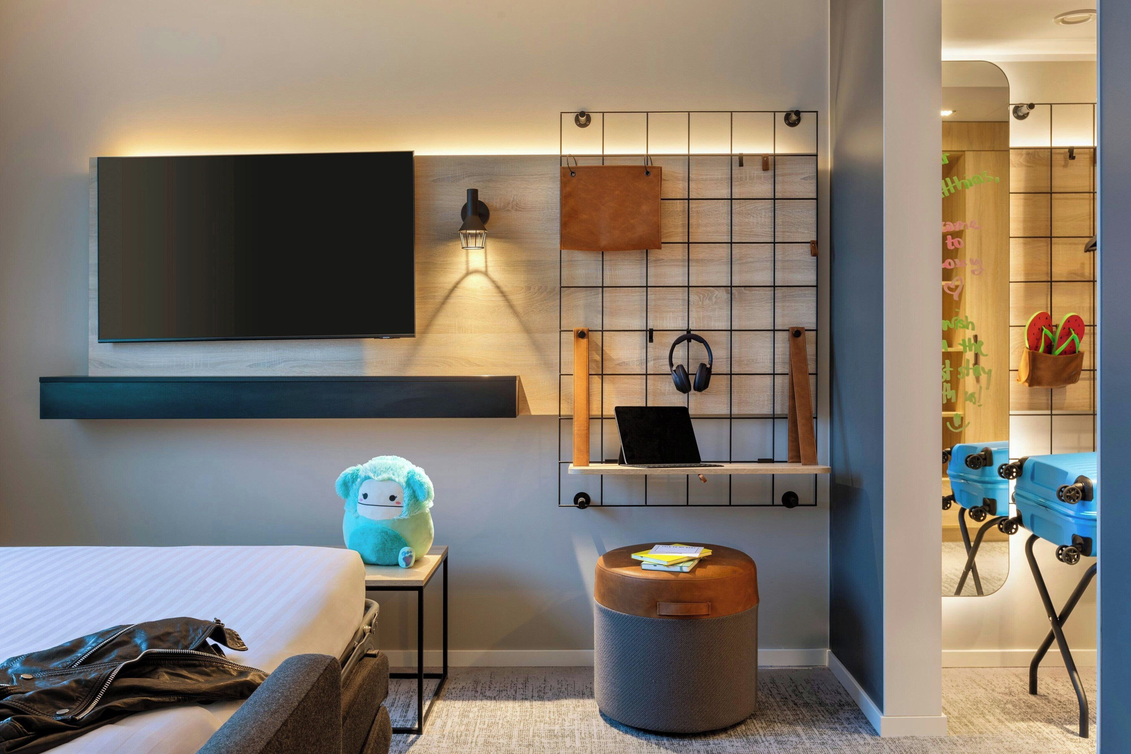 A modern hotel room with a large flat-screen TV, a wall-mounted shelf with a laptop, headphones, and a small plush toy. There is a grey ottoman with a note on it, and a suitcase rack with two suitcases.