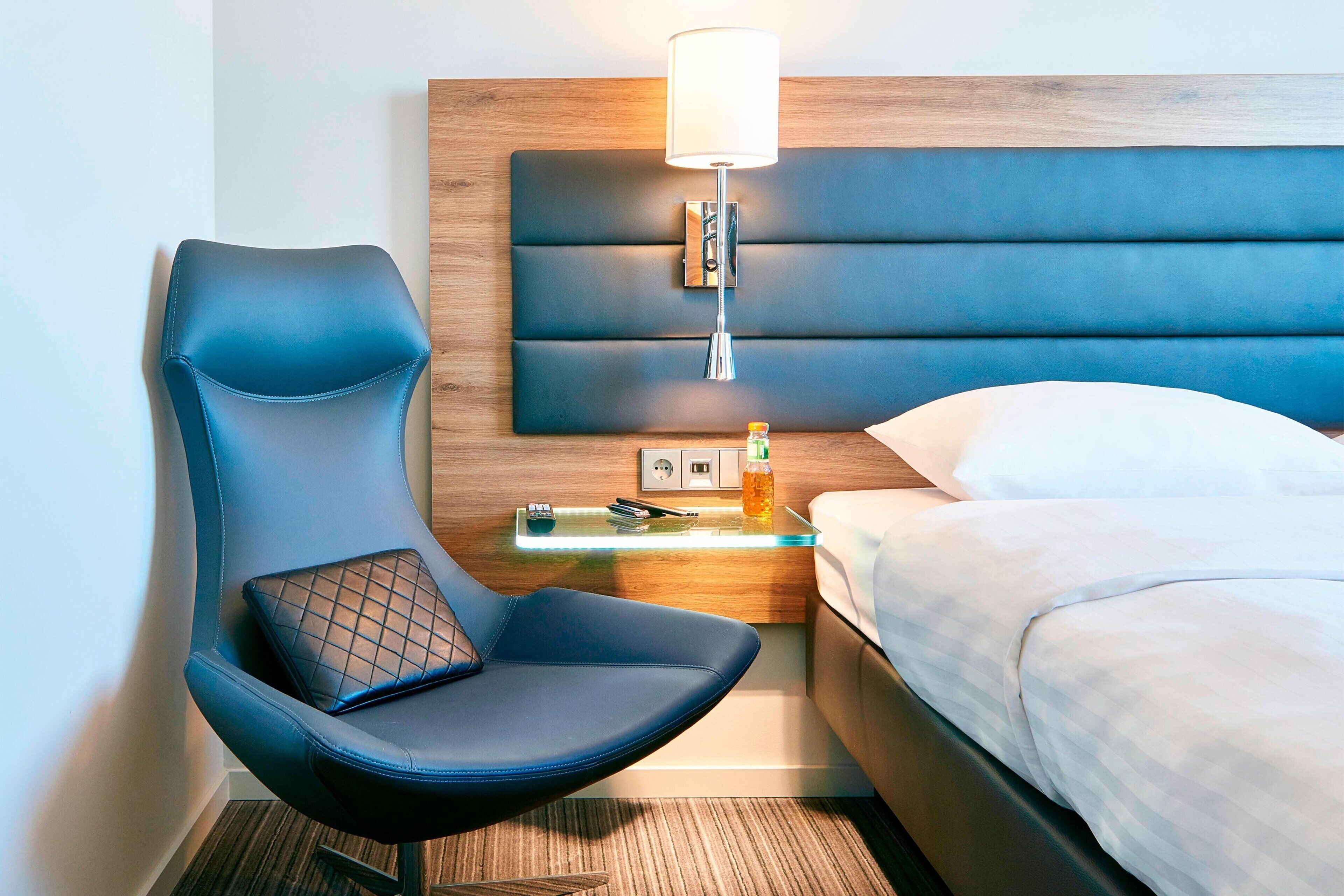A modern hotel room with a blue upholstered chair, a bedside table with a lamp, and a neatly made bed.
