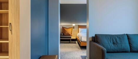 A modern hotel room with a grey sofa, a wooden wardrobe, a small seating area with cushions, and a bed with a wooden headboard.