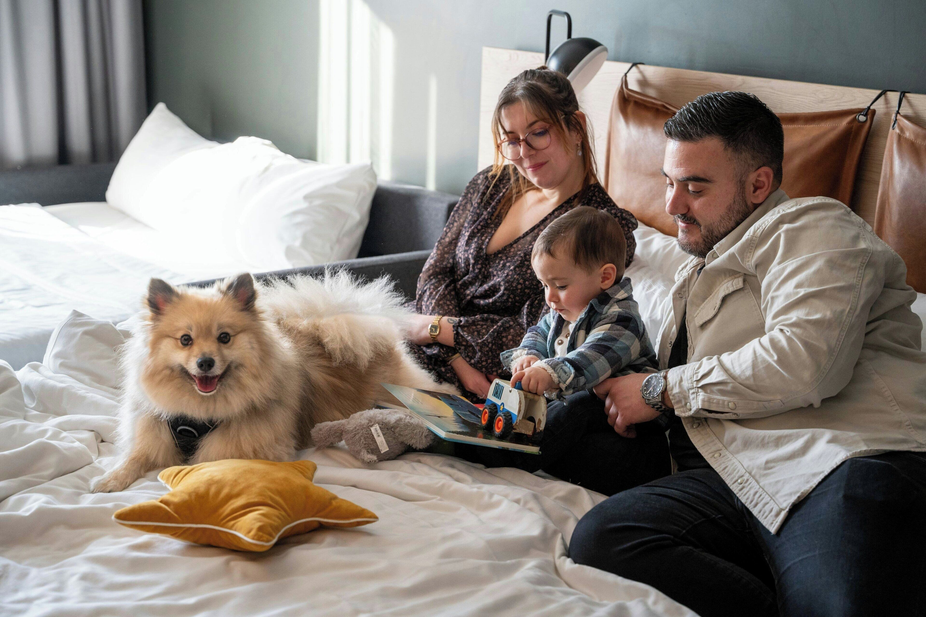 Una famiglia con un cane su un letto con lenzuola bianche, un cuscino giallo e un libro.