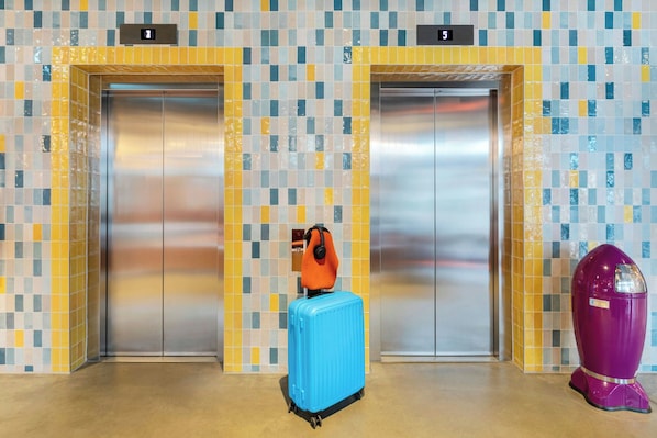 Un ascensor con pared de azulejos, una maleta azul y un objeto morado.