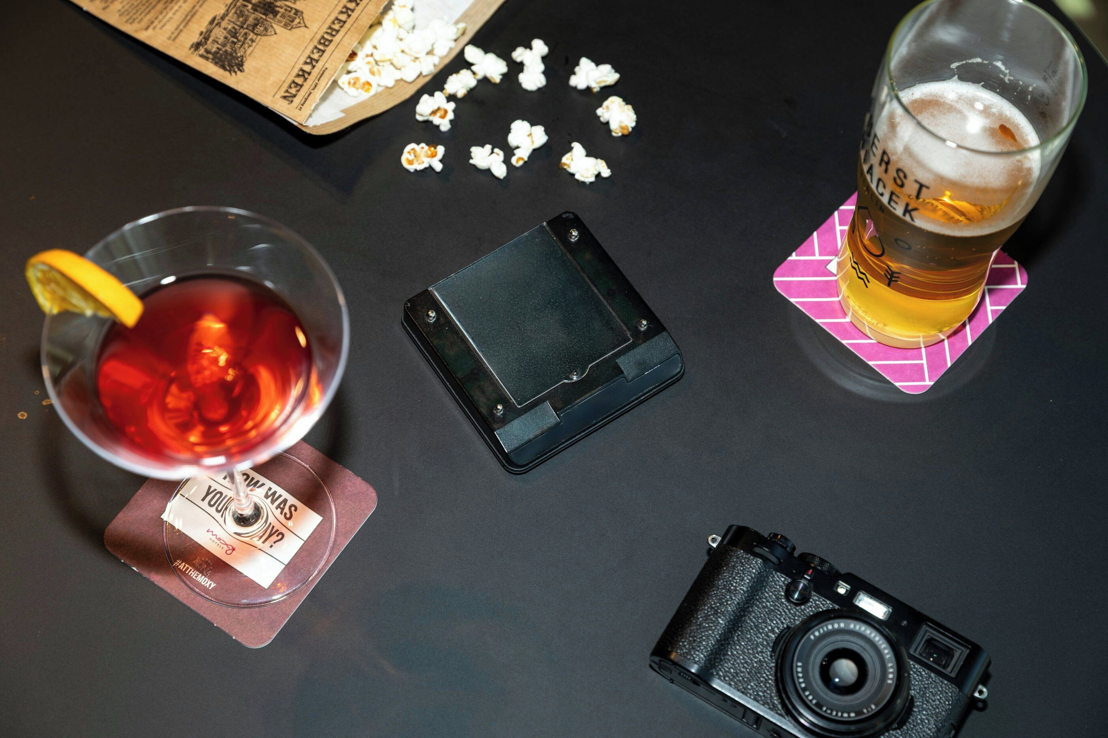 A table with a glass of beer, a camera, a smartphone, and scattered popcorn.
