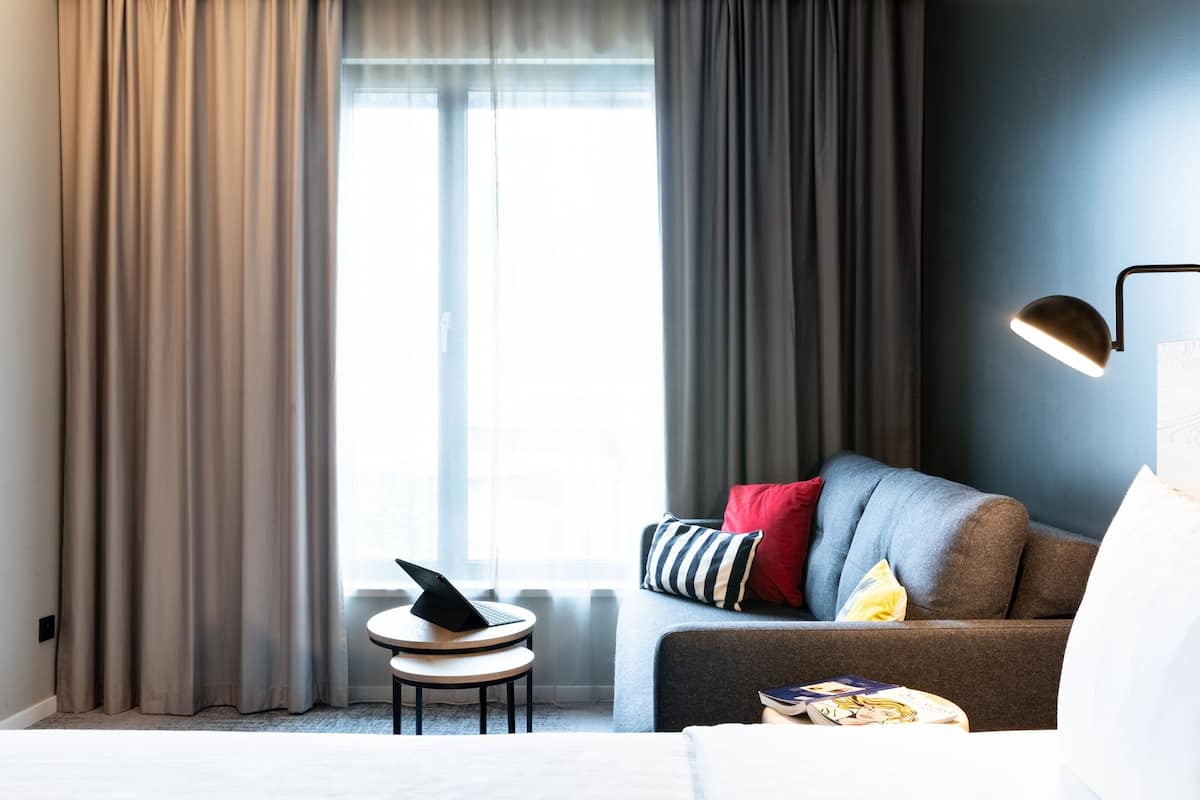 A modern hotel room with a bed, a round table with a laptop, a sofa with cushions, and a lamp.