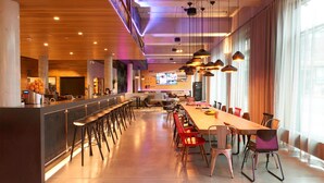 A modern restaurant with a long counter, multiple bar stools, and a mix of red and white chairs. - Moxy Berlin Ostbahnhof (Berlin)