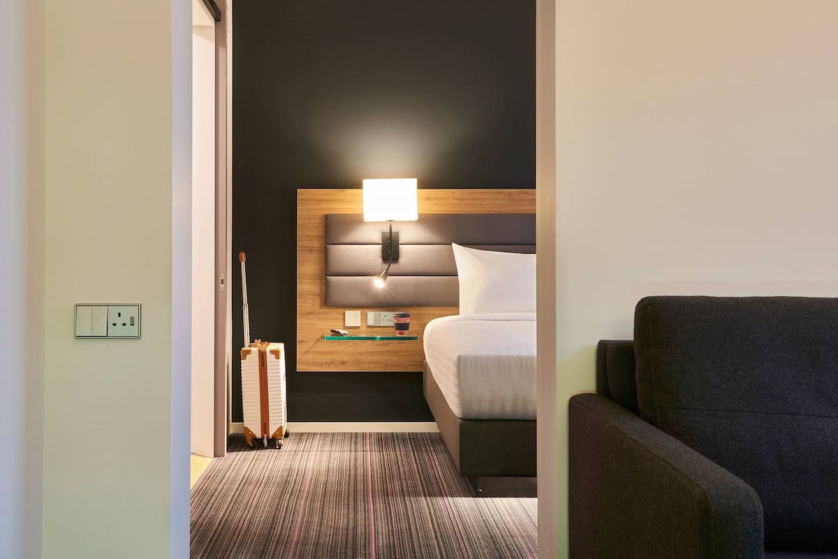 A hotel room with a bed, a bedside table with a lamp, a chair, and a carpeted floor.