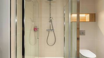 A modern bathroom with a glass-enclosed shower, a white toilet, and a wooden shelf above the toilet.
