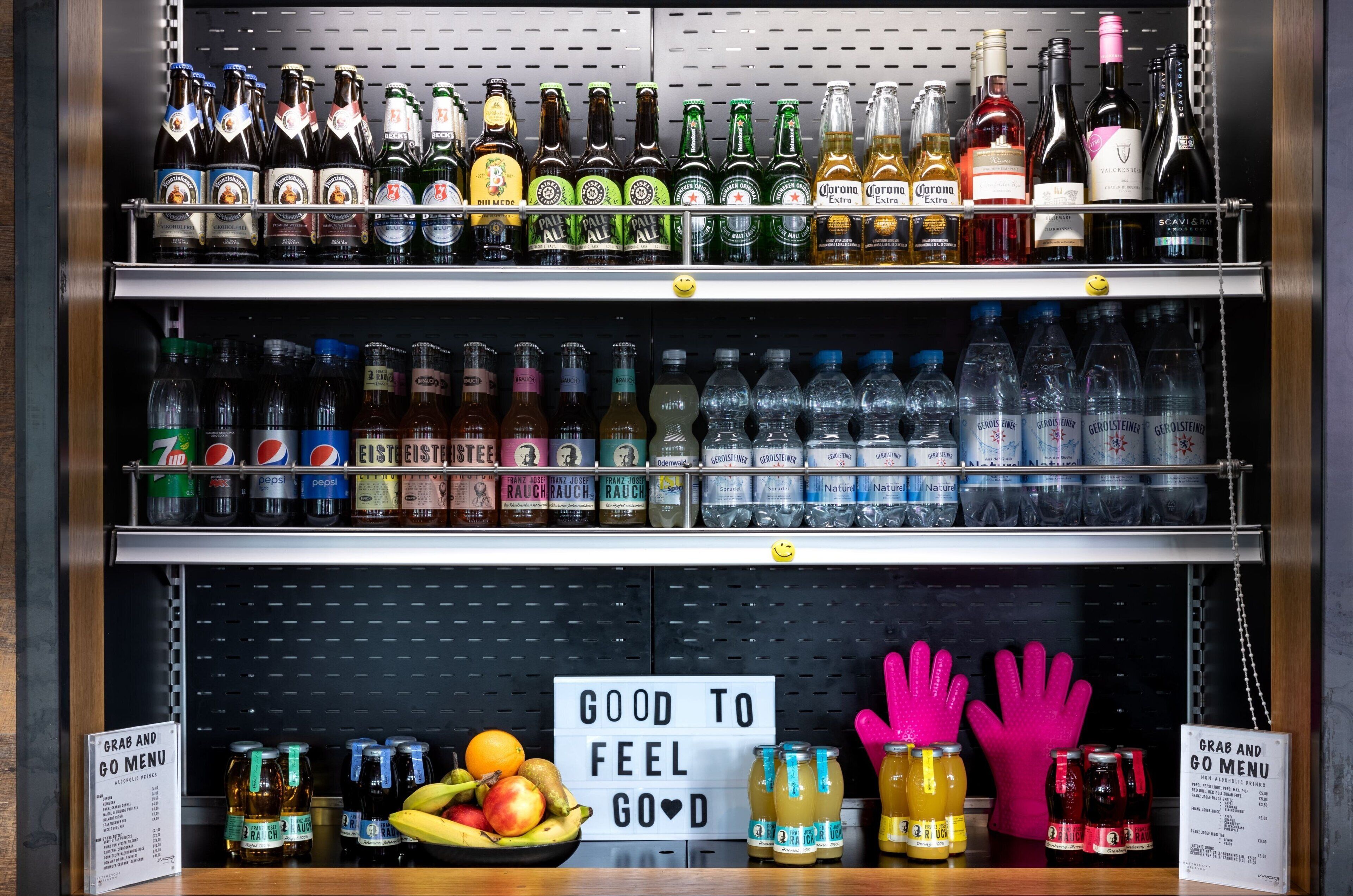 A shelf stocked with various beverages including beer, wine, and soda. Below the shelf, there is a sign that reads "GOOD TO FEEL GOOD" and a display of fruits and bottles.