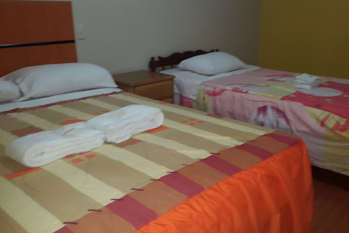 Habitación de hotel con dos camas, cada una con un edredón de color diferente. Hay una mesita de noche de madera entre las camas, y una toalla blanca está colocada en la cama en primer plano.