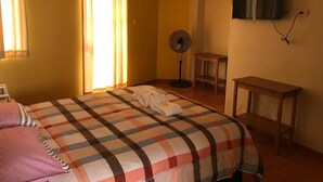 A hotel room with a large bed, a TV mounted on the wall, a fan, and a small table.