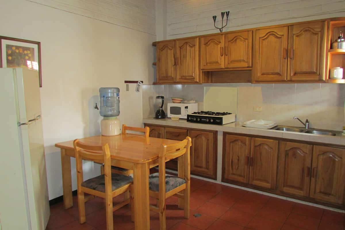 A compact kitchen with wooden cabinets, a dining table with chairs, a refrigerator, a water dispenser, a microwave, a stove, and a sink.