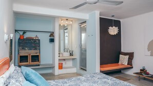 Une chambre à coucher comprenant un lit, un banc, un ventilateur de plafond, une commode, un miroir et une porte donnant sur une salle de bain.