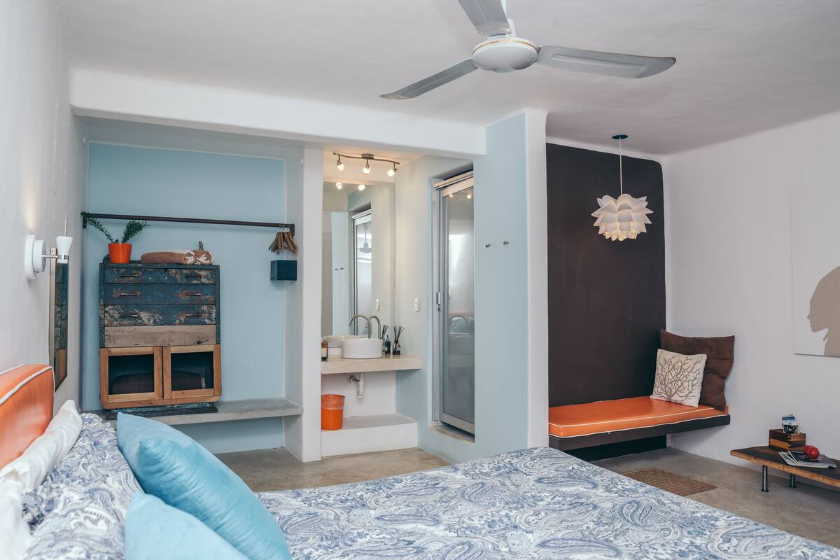 A bedroom with a bed, a bench, a ceiling fan, a dresser, a mirror, and a door leading to a bathroom.