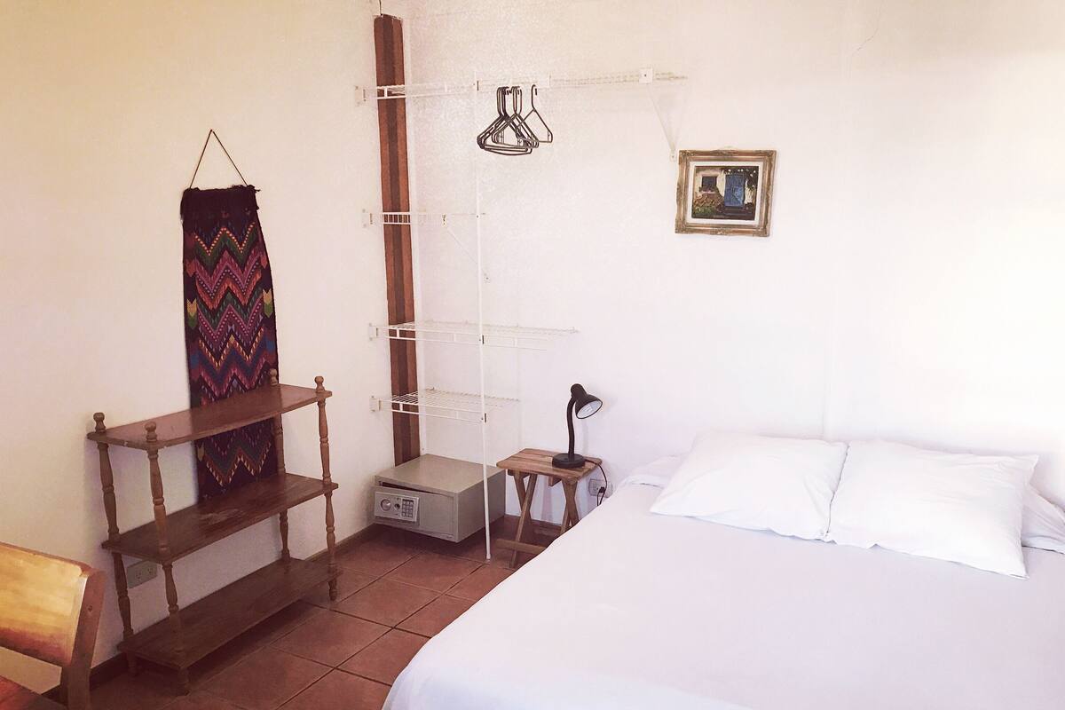 A bedroom with a bed, a desk, a chair, a shelf, a lamp, and a wall-mounted rack.