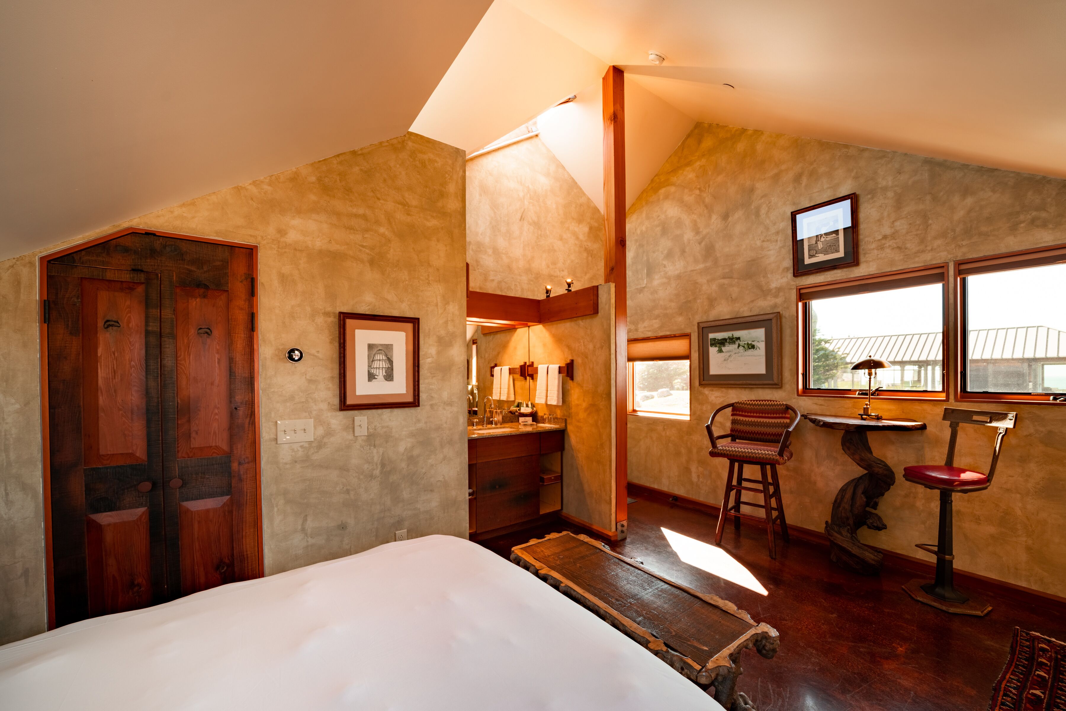 A room with a bed, wooden door, framed pictures, a chair, a table, and a window.