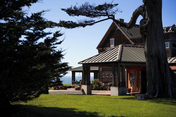 A large wooden house with a covered patio, a large tree, and a view of the ocean.
