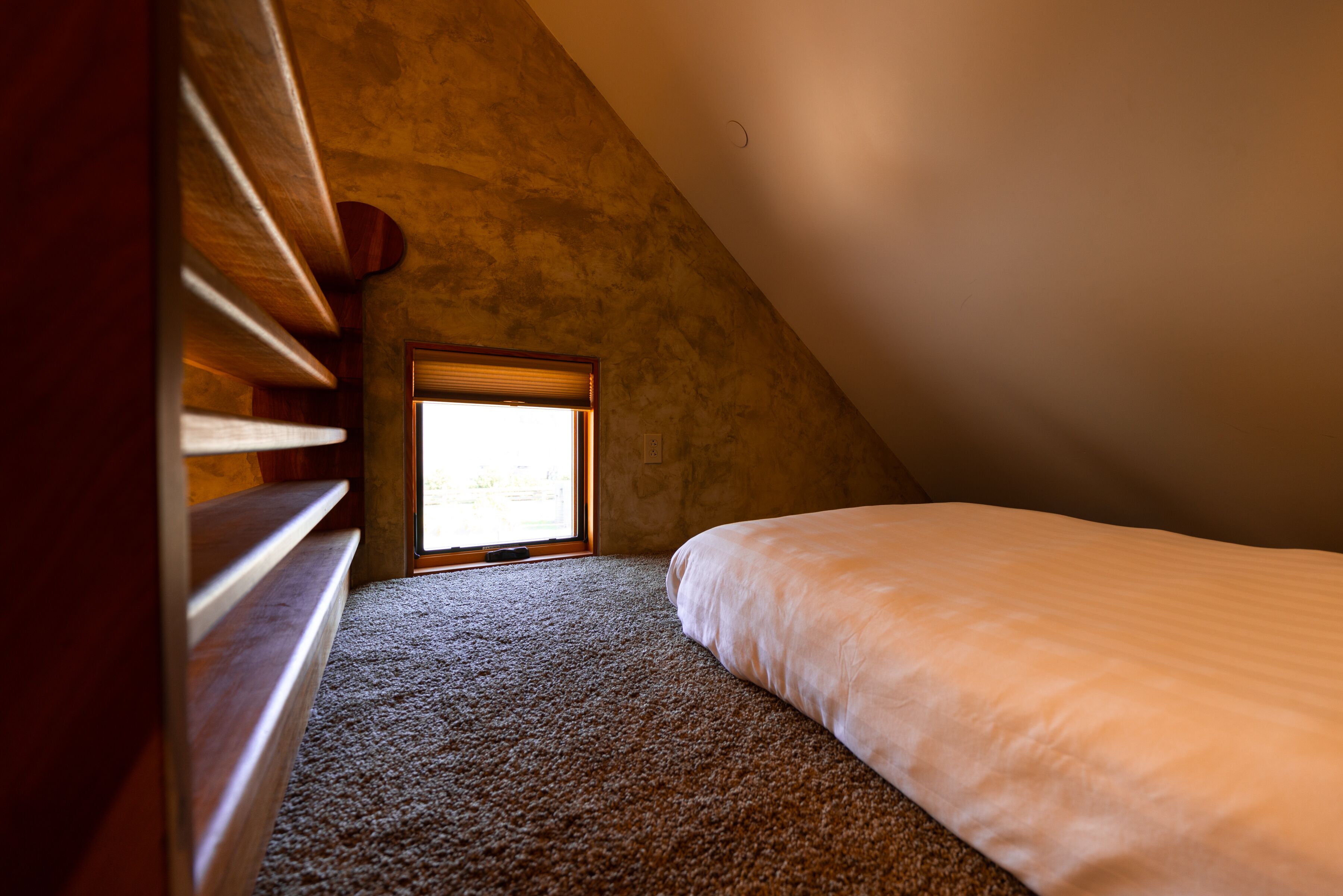 A bedroom with a sloped ceiling, a window with blinds, a bed, and a wooden bench.