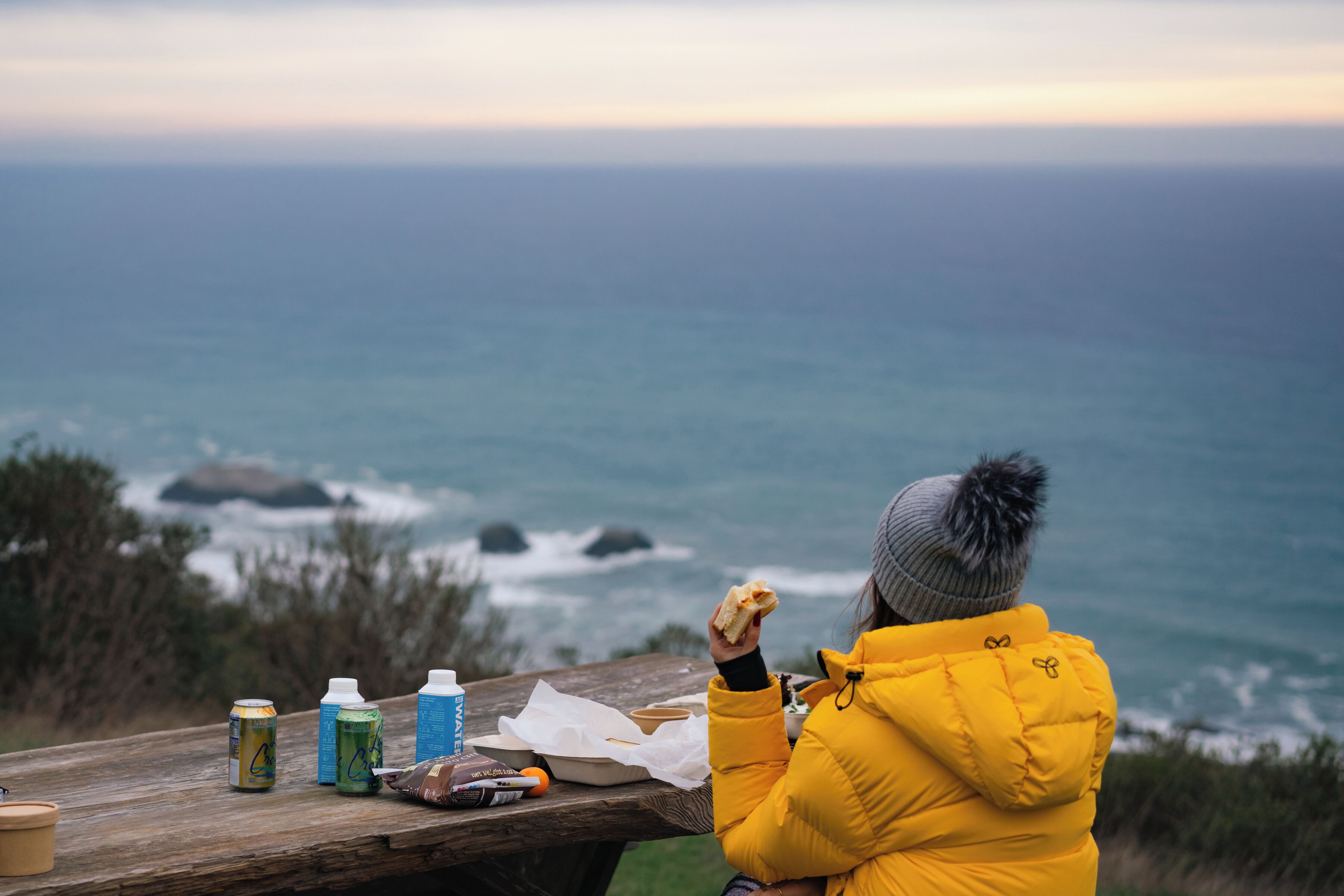 Eine Person in einer gelben Jacke sitzt an einem Holztisch mit Blick auf das Meer.