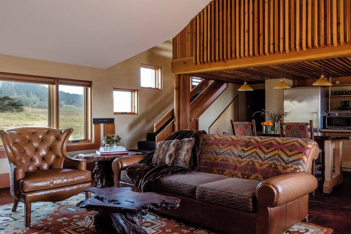 Amplia sala de estar con un sillón de cuero, un sofá estampado, una mesa de centro de madera y vistas al exterior.